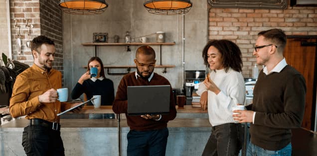 Group of coworkers in an office kitchen collaborating around a laptop while chatting and holding coffee.